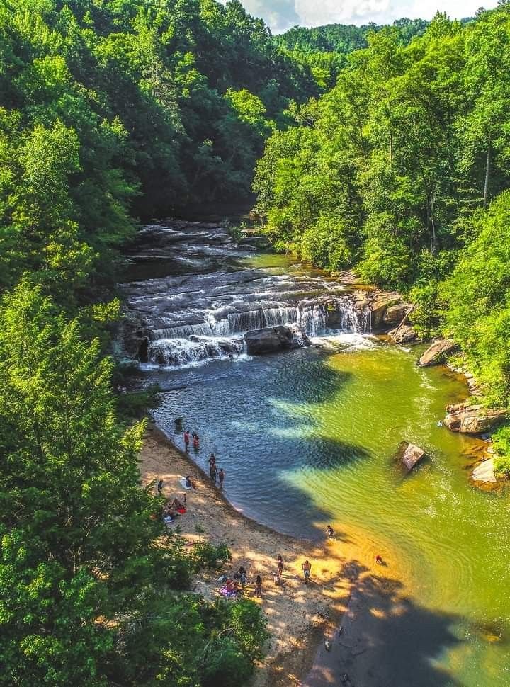 This may contain: people are standing at the edge of a river with waterfall in the background and green trees surrounding it