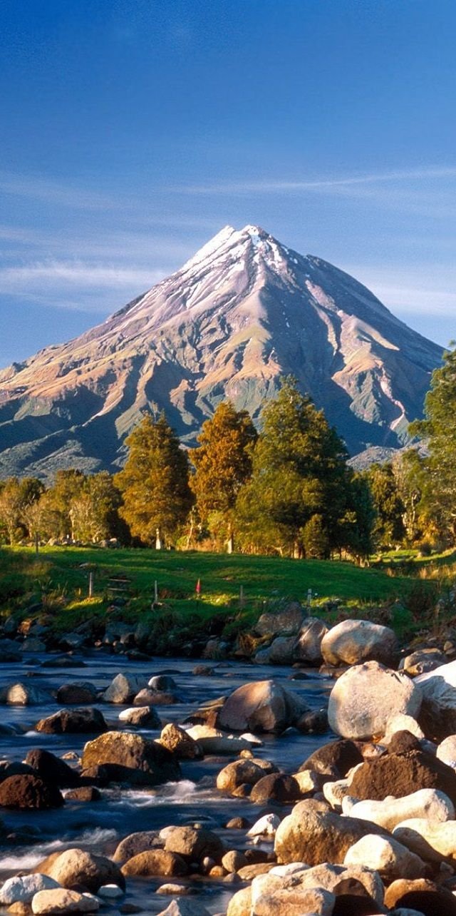 This may contain: there is a mountain in the background and rocks on the river bank, with trees around it