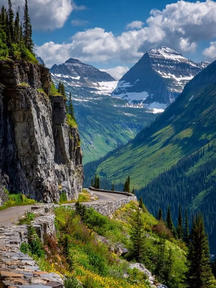 This may contain: a scenic road in the mountains with trees and flowers on both sides, surrounded by snow capped mountains