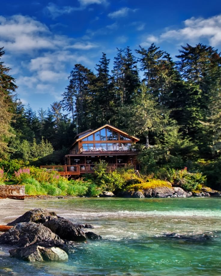 This may contain: a house on the shore of a river surrounded by trees and rocks in front of it
