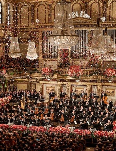 This may contain: an orchestra performing in front of a large crowd with christmas decorations on the ceiling and chandelier