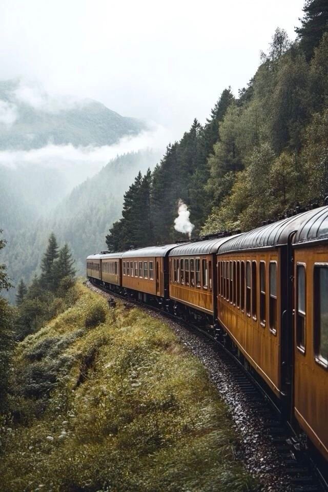This may contain: a train traveling down tracks next to a lush green hillside covered in fog and low lying clouds
