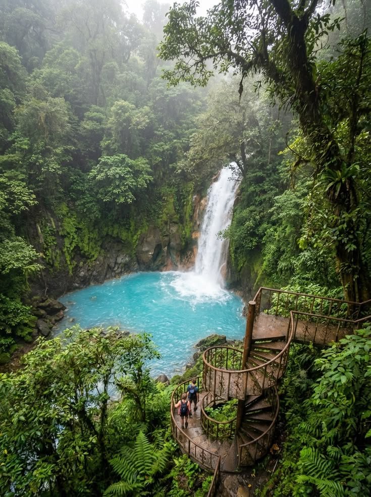 This contains: Aerial view of Rio Celeste waterfall in Costa Rica cascading into a vivid electric turquoise blue pool, surrounded by dense misty rainforest, two hikers with backpacks descending a rustic iron spiral staircase in the foreground, lush ferns and jungle vegetation, soft foggy light filtering through the canopy.