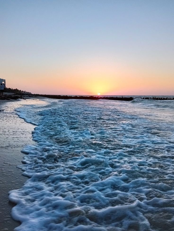 This may contain: the sun is setting over the ocean with waves coming in to shore and buildings on the other side