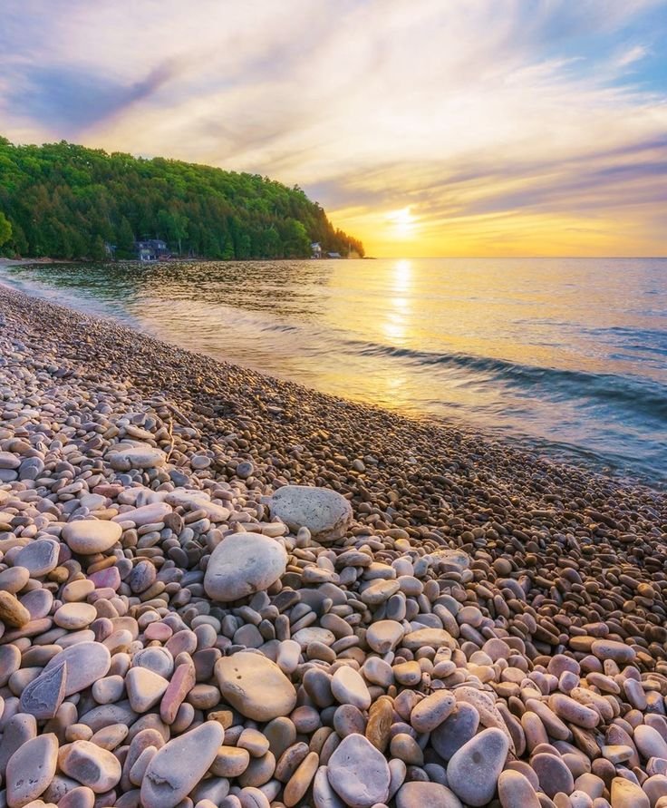 This may contain: the sun is setting over the water and some rocks on the beach with trees in the background