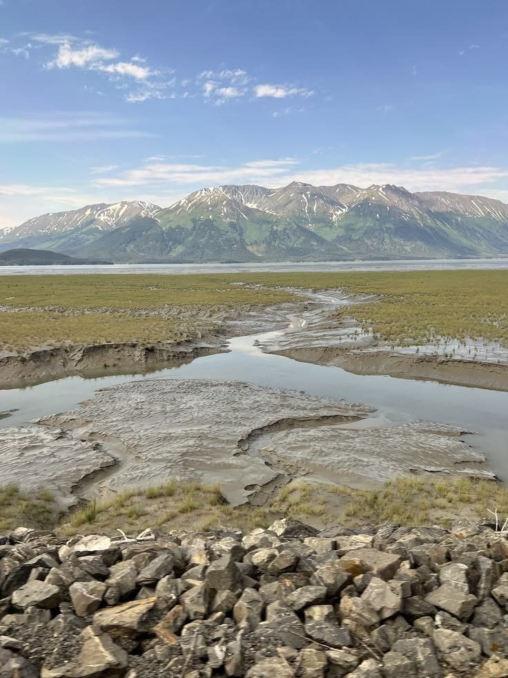 This may contain: there is a mountain range in the distance with water running through it and rocks on the ground