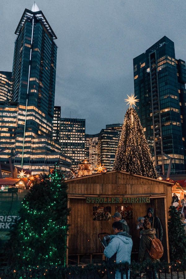 This may contain: people are standing in front of a small christmas tree with lights and buildings behind it