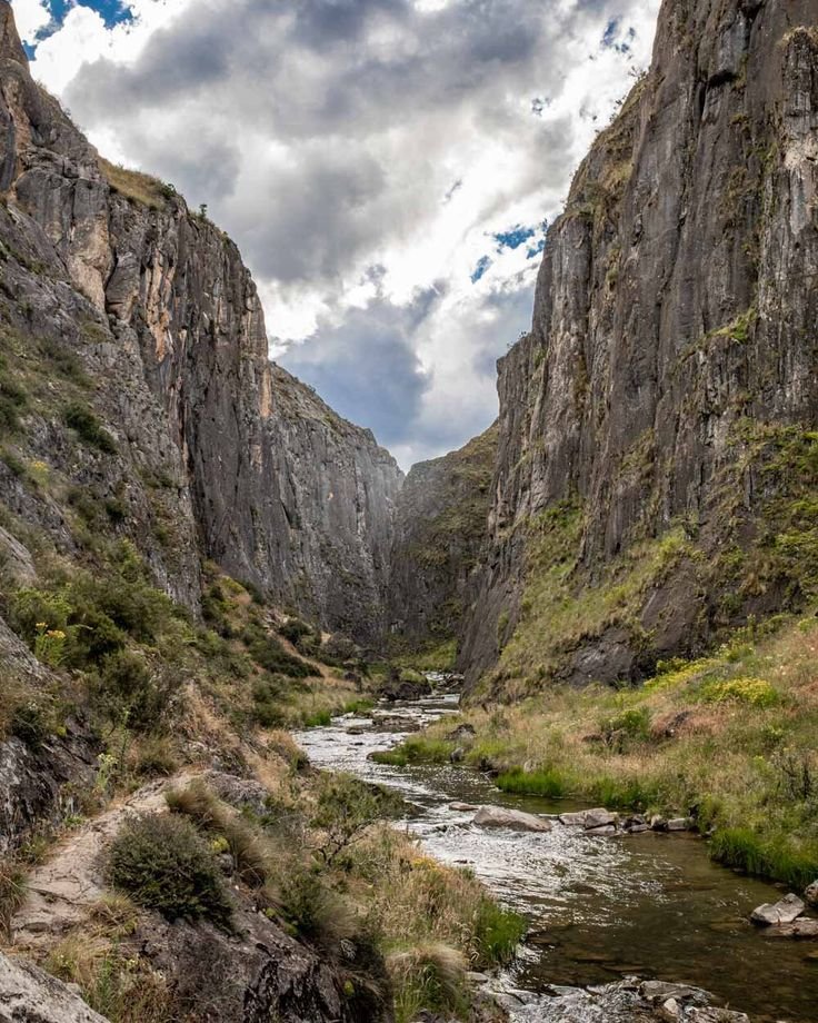 This may contain: a river flowing through a canyon surrounded by tall rocky mountains under a cloudy blue sky