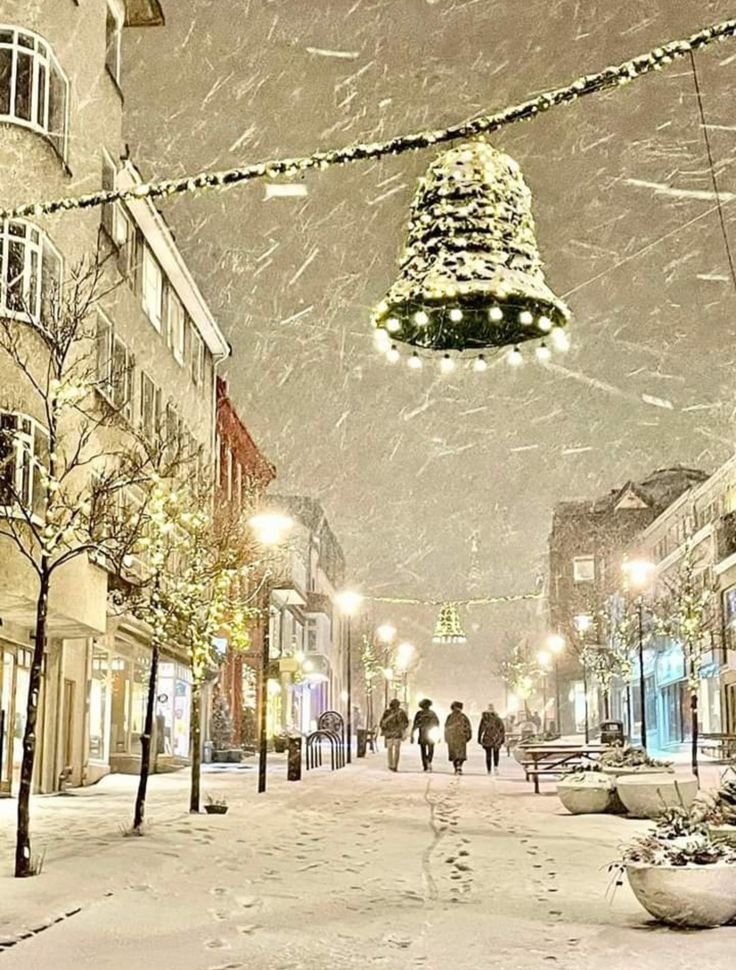 This may contain: people walking down a snowy street at night with christmas lights hanging from the ceiling above them