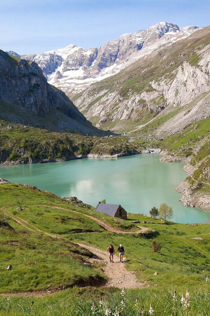This may contain: two people walking up a trail towards a lake with mountains in the backgroud