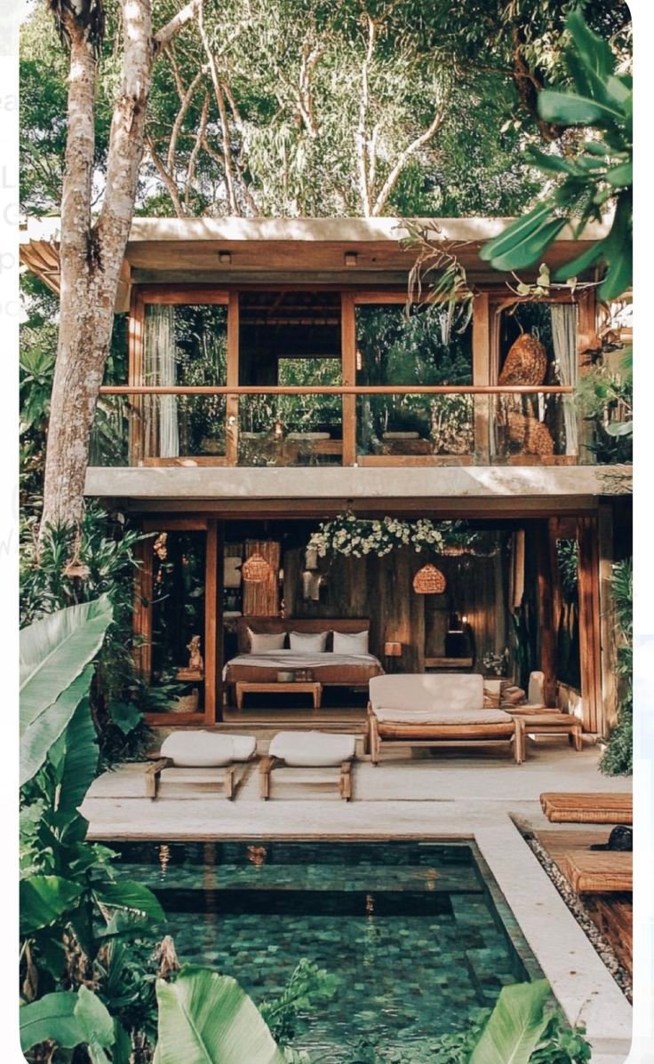 This may contain: an outdoor pool surrounded by greenery and wooden furniture in front of a large house