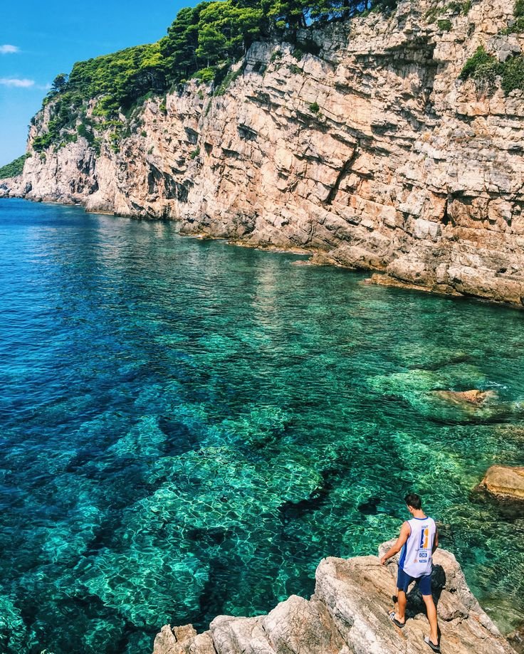 This may contain: a man sitting on top of a rock next to the ocean with clear blue water